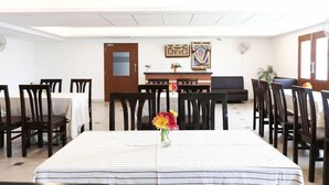 A dining area with a long table, multiple chairs, and a small vase with flowers. There is a wooden door and a framed picture on the wall. - Treebo Akshaya Mahal Inn (Mysore)