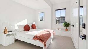 Un dormitorio moderno con una cama blanca, almohadas rojas, una mesita de noche con lámpara, un ventanal con una planta y una puerta con cerradura electrónica.