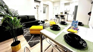 Ein modernes Wohnzimmer mit einer schwarzen Couch, einem gelben Hocker und einem Essbereich mit schwarzen Tellern und Wasserflaschen.