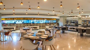 A spacious restaurant with high ceilings and pendant lighting. Tables are set with white tablecloths and various dishes. A bar area with a patterned backsplash is visible in the background. - Zone By The Park Jodhpur (Jodhpur)
