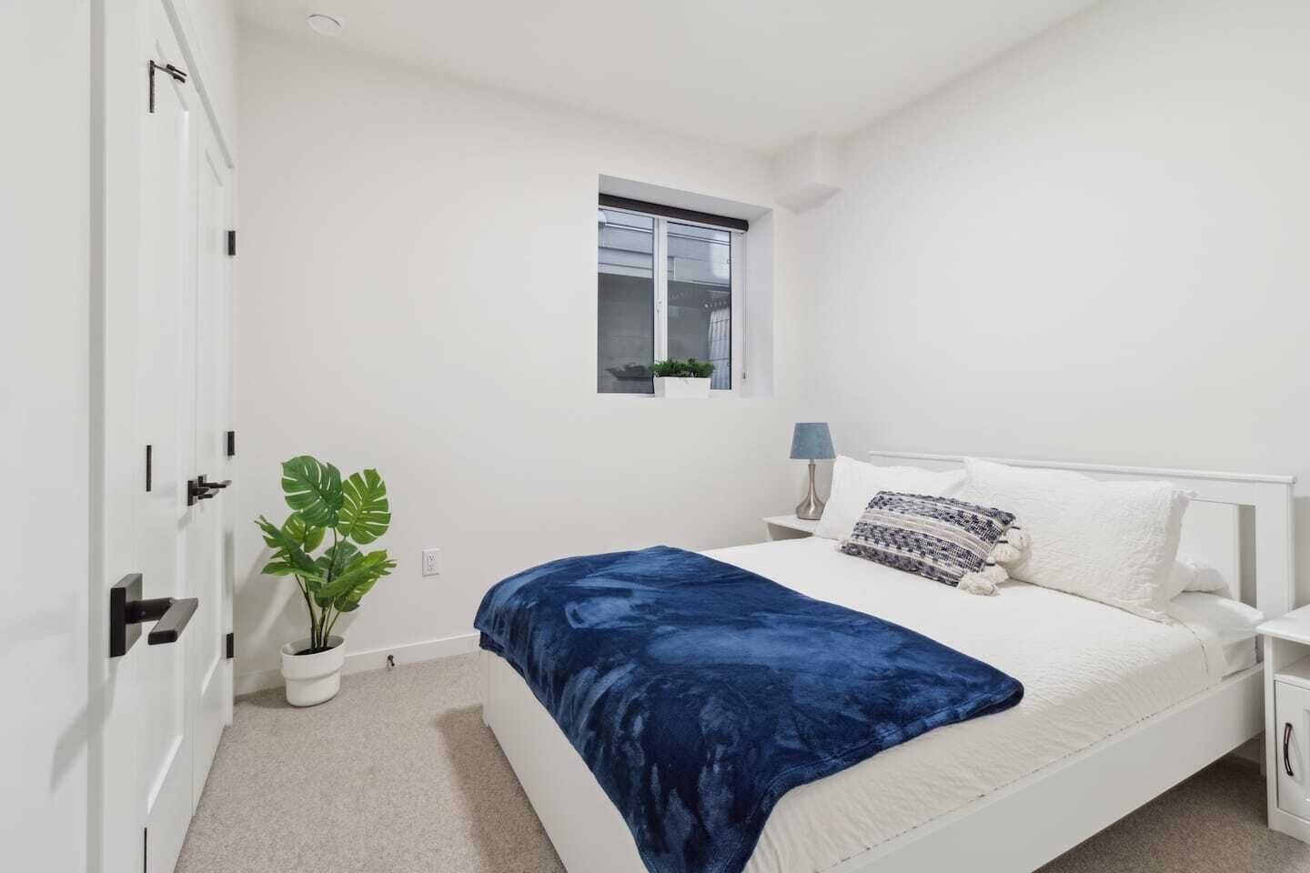 Un dormitorio con cama, ventana, mesita de noche, lámpara y una planta en maceta.