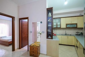 Quarto com cama, porta de madeira, pequeno baú de madeira, cozinha com armários e pia, e espelho com prateleira acima.