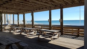 Una terraza de madera con mesas de picnic y bancos que da a una playa.