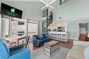 A modern living room with a wooden table, blue armchairs, a vintage trunk, and a kitchen area with white cabinets and a spiral staircase. - Top Floor Gulf Views | Private Beach + Free Chairs | Sleeps 10 | Coral Reef Club 49 (Destin)