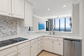 A modern kitchen with white cabinetry, a black stovetop, a stainless steel dishwasher, and a large window offering a view of the ocean. - Beachfront Penthouse~Breath Taking Views~3 King Suites~2 Balconies! (Destin)