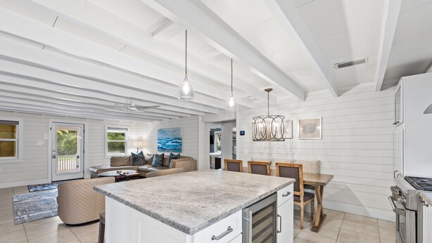 Cozinha espaçosa com uma ilha grande, armários brancos e eletrodomésticos de aço inoxidável. Há uma sala de jantar com mesa e cadeiras, e uma sala de estar com sofá e um quadro na parede.
