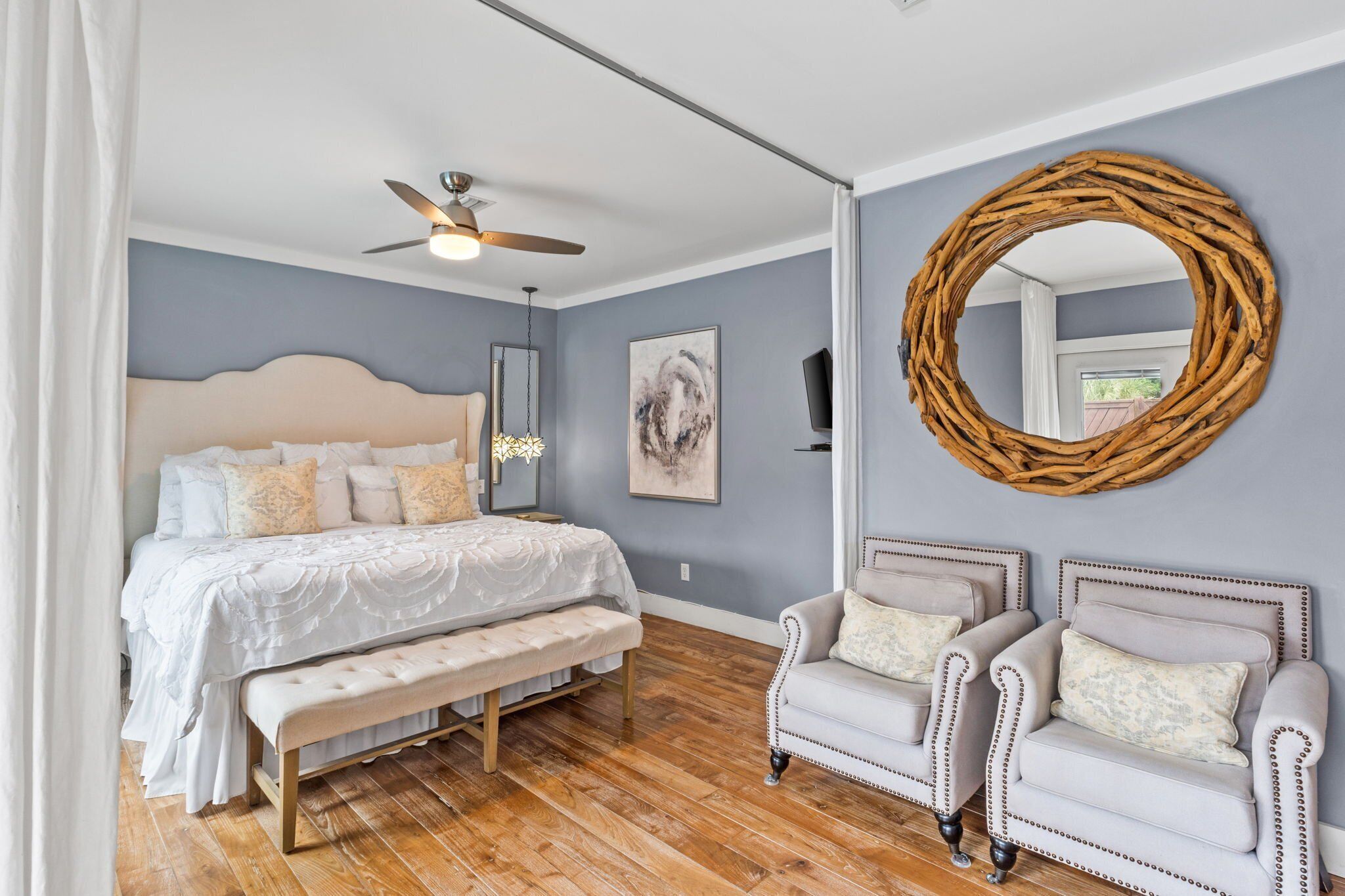 Une chambre à coucher comprenant un lit, un banc, deux fauteuils, un ventilateur de plafond, un miroir et une œuvre d’art encadrée accrochée au mur.