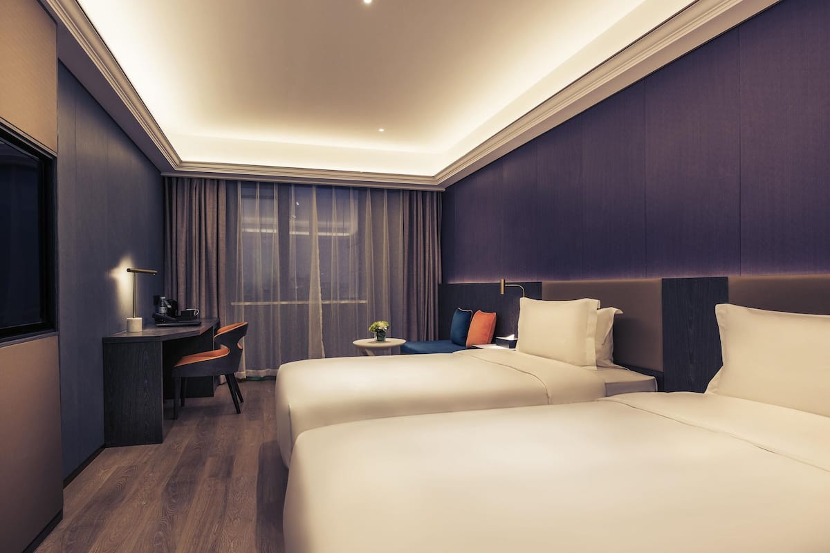 Habitación de hotel con dos camas, un escritorio, una silla, una mesita con un florero y una ventana con cortinas.