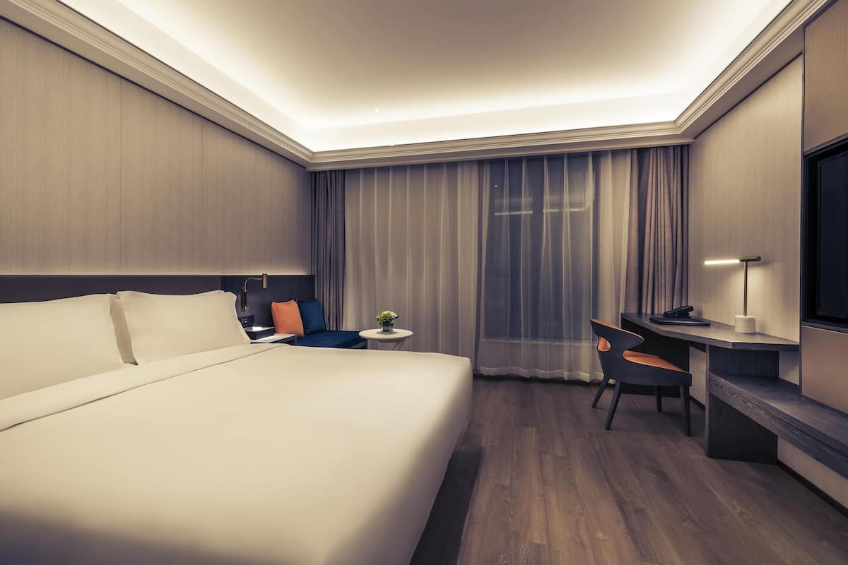Una habitación de hotel con una cama grande, un escritorio, una silla, una mesita con una planta y un televisor en la pared.