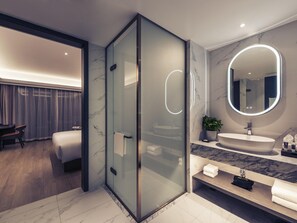 Une salle de bain moderne dotée d’une douche à parois de verre, d’un meuble-lavabo recouvert de carreaux de marbre et d’un grand miroir avec éclairage intégré.