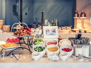 Un buffet proposant une variété d’aliments, notamment des fruits, des légumes et des condiments, disposés sur une table agrémentée d’une reproduction décorative de la Tour Eiffel et d’un tableau encadré.