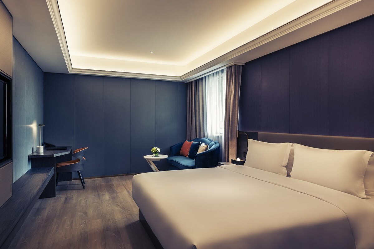 A modern hotel room with a large bed, a desk, a chair, and a small table with a vase of flowers.