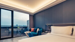 A hotel room with a large bed, a desk with a chair, a blue armchair, and a view through a glass door.