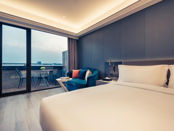 Habitación de hotel con una cama grande, un escritorio con una silla, un sillón azul y vistas a través de un ventanal.