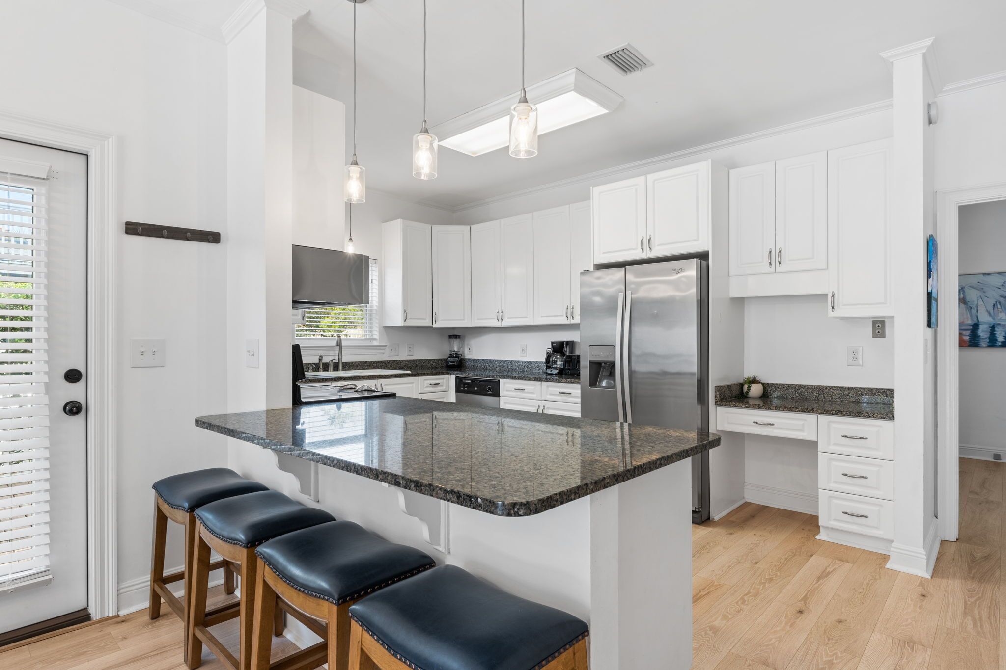 A modern kitchen with white cabinets, a granite countertop, and a stainless steel refrigerator. There are three stools at the island and pendant lights above.
