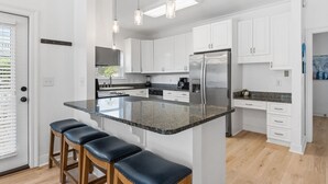 Una cocina moderna con armarios blancos, encimera de granito y refrigerador de acero inoxidable. Hay tres taburetes en la isla y luces colgantes arriba.