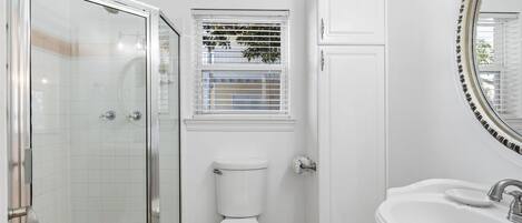 Une salle de bain avec une douche à parois de verre, des toilettes, une fenêtre avec des stores et un lavabo avec un miroir.