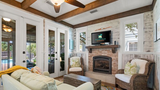 A cozy living room with a fireplace, wicker chairs, a sofa, and a television mounted above the fireplace. - Private Pool, Steps to Beach, Screen in Porch, Cutest Cottage in Crystal Beach! (Destin)