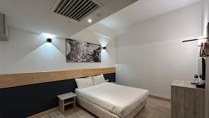 Une chambre à coucher comprenant un lit, une petite table de chevet, une tête de lit, une œuvre d’art accrochée au mur et une bouche d’aération au plafond.