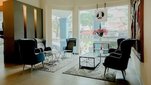 Un hall d’hôtel moderne avec une table basse en verre, des chaises noires et bleues, un vase rempli de fleurs roses et de grandes fenêtres donnant sur la rue.