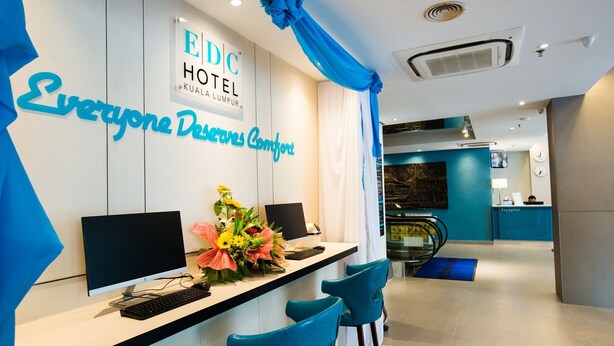 Un comptoir d’accueil avec un ordinateur, un arrangement floral et une enseigne indiquant « EDC HOTEL KUALA LUMPUR ».