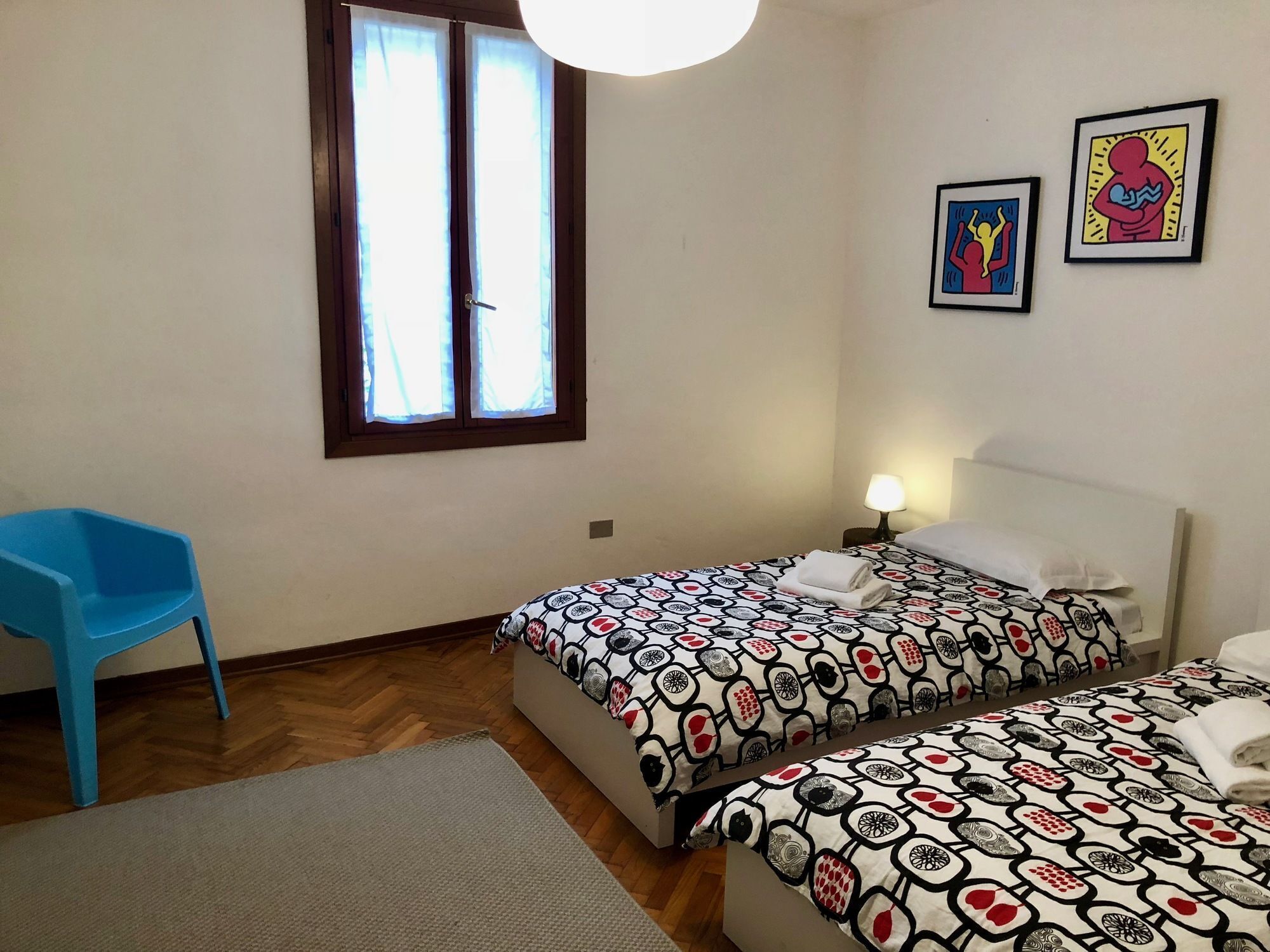 Una habitación con dos camas, una silla azul, una ventana con cortinas y dos cuadros en la pared.