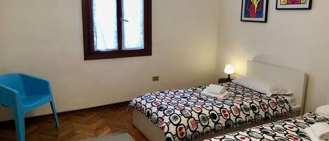 Una habitación con dos camas, una silla azul, una ventana con cortinas y dos cuadros en la pared.