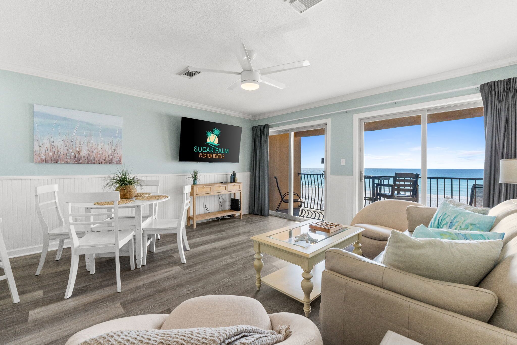 A spacious living room with a balcony overlooking the ocean, a dining area with white furniture, and a flat-screen TV displaying 'Sugar Palm Vacation Rentals'.