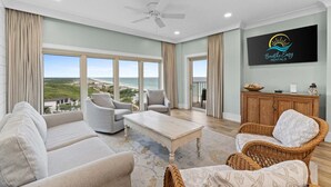 A spacious living room with a large window, a ceiling fan, and a flat-screen TV mounted on the wall. - New! Beach Manor 1005 Gorgeous Remodeled Condo w/ Gulf Views & Resort Amenities (Miramar Beach)