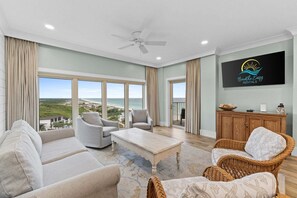 A spacious living room with a large window, a ceiling fan, and a flat-screen TV mounted on the wall. - New! Beach Manor 1005 Gorgeous Remodeled Condo w/ Gulf Views & Resort Amenities (Miramar Beach)