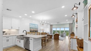 A modern kitchen with white cabinetry, a central island, and a dining area with wicker chairs. - New! Beach Manor 1005 Gorgeous Remodeled Condo w/ Gulf Views & Resort Amenities (Miramar Beach)