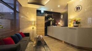 A modern hotel lobby with a reception desk, a sofa with colorful cushions, a coffee table, and a wall clock. - Guseo Station Brown Dot (Busan)