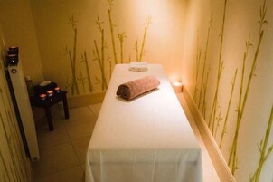 Un cuarto de spa con una camilla de masaje, una toalla, velas y una mesita con una botella y un recipiente.