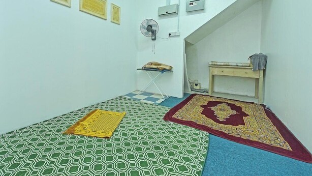 Une petite pièce avec un sol à carreaux, un lit recouvert d’une courtepointe à motifs, un ventilateur et une petite table avec un coussin.