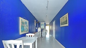 Un couloir aux murs bleus, au plafond blanc et au sol recouvert de carreaux blancs. Des tableaux encadrés ornent les murs, et une table avec des chaises se trouve au fond du couloir.