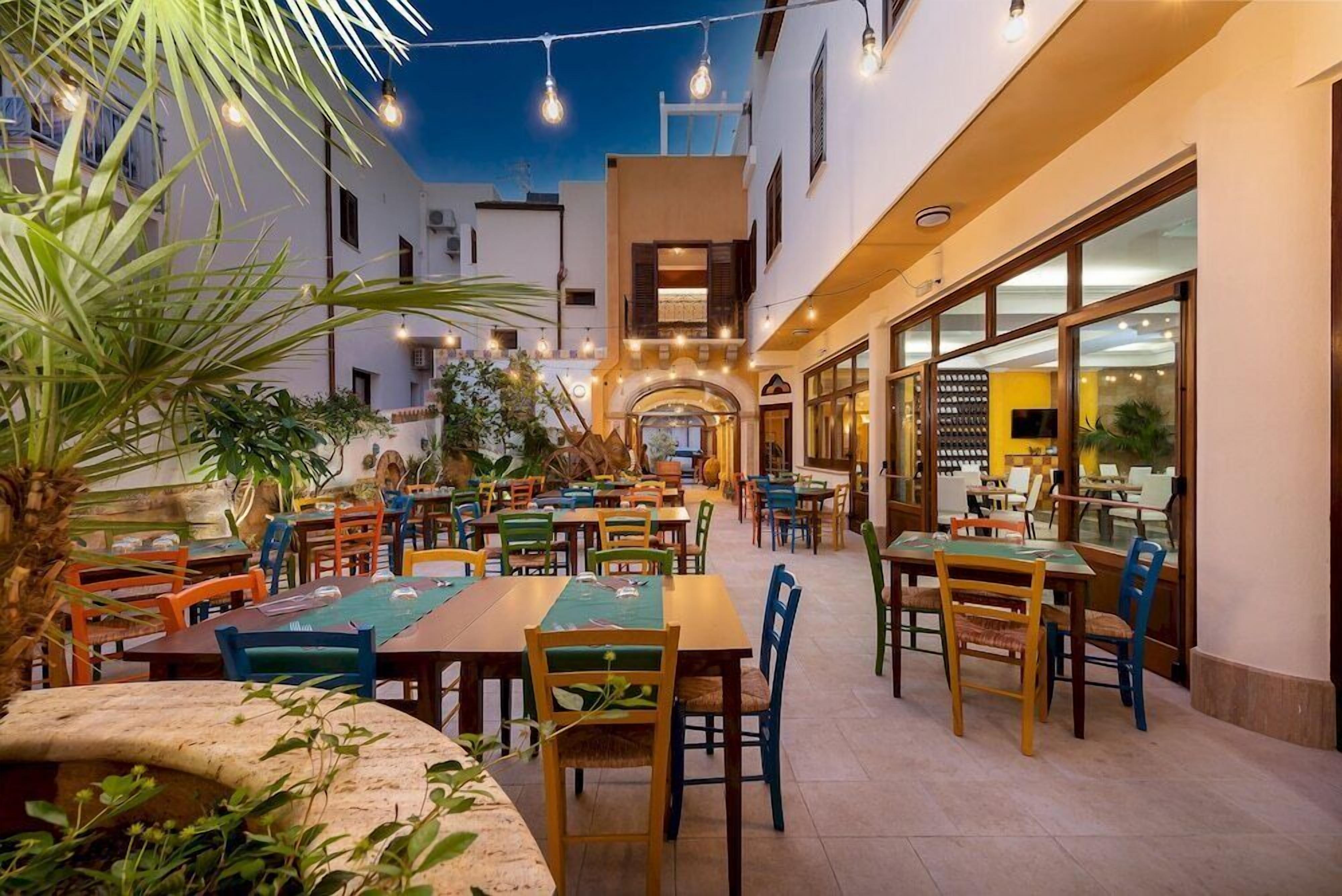 An outdoor dining area with multiple tables and chairs, surrounded by plants and a building with large windows.