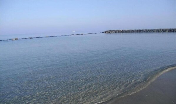 A calm beach with clear water, a rocky breakwater, and a distant shoreline.
