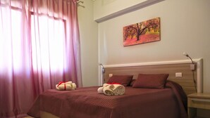 A neatly made bed with a red bedspread and pillows, a painting on the wall, and a window with pink curtains.