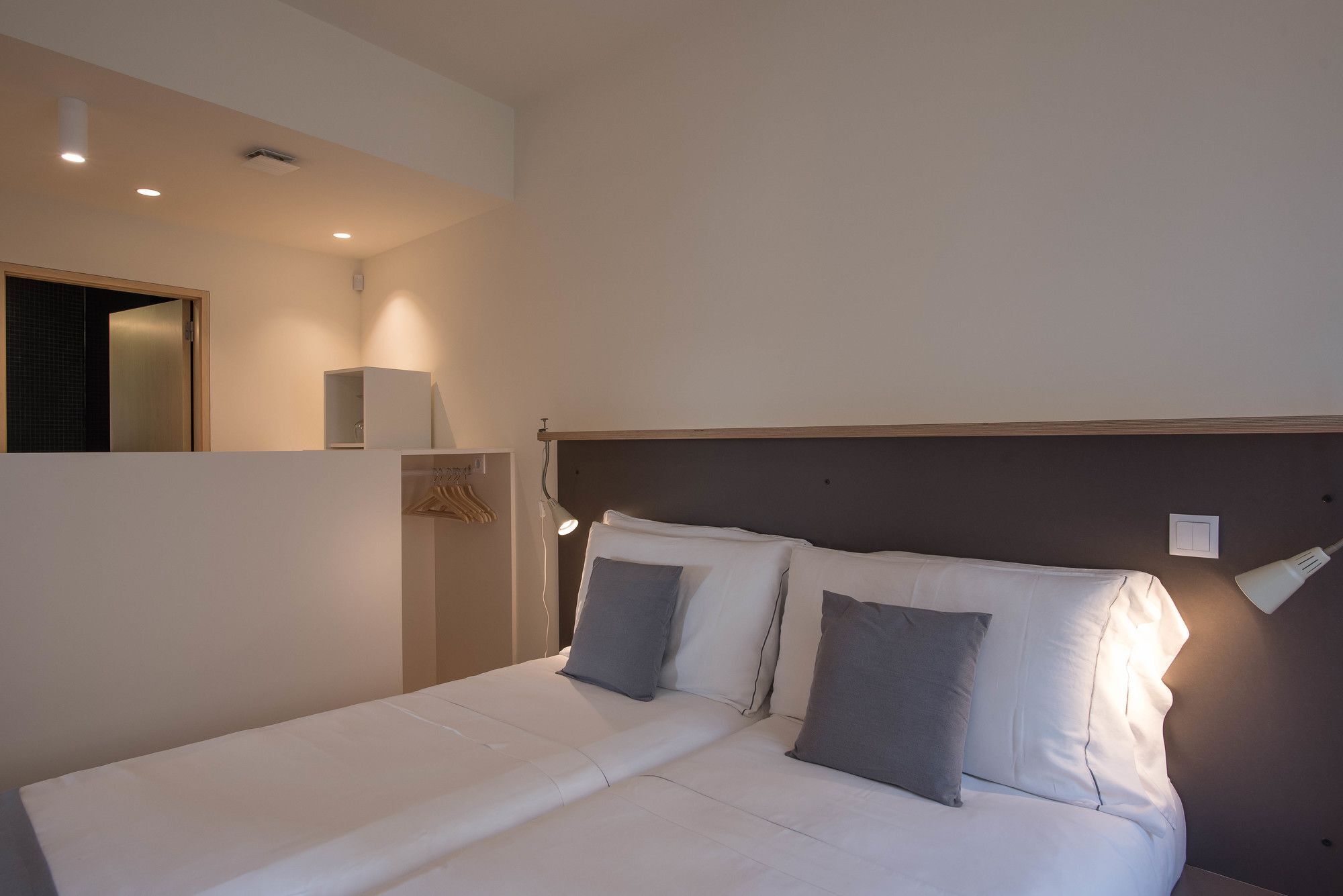 Una cama bien tendida con sábanas blancas y dos almohadas, una lámpara de noche y un pequeño estante sobre la cama.
