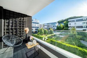 A balcony with a modern chair, a small table with a vase and cups, and a view of a well-maintained garden and buildings in the background. - Relax in Gdynia's heart - comfortable & beach-close (Gdynia)