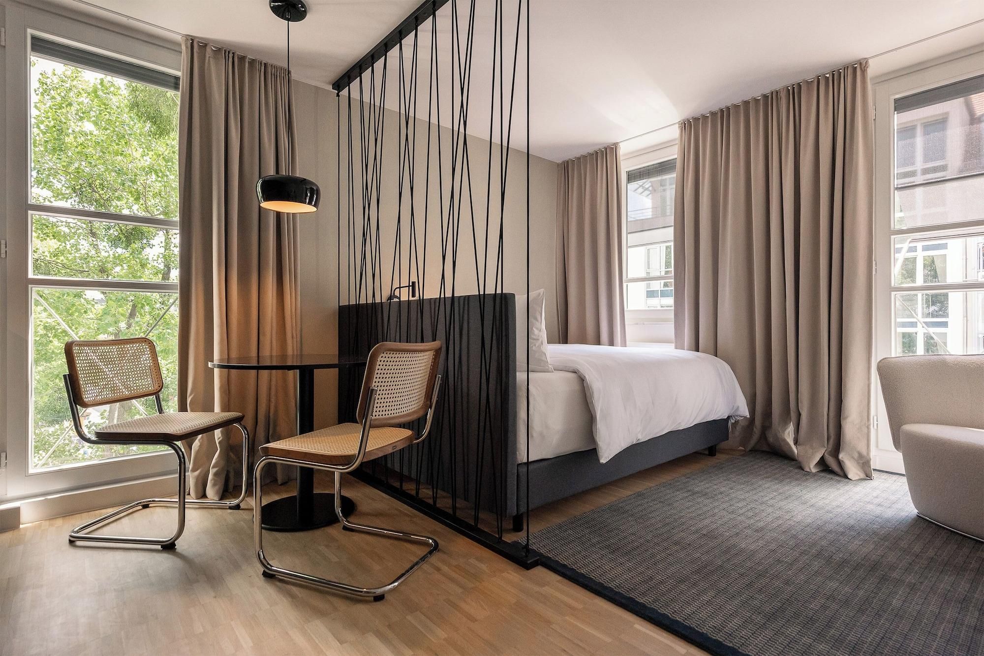 A modern hotel room with a bed, two chairs, a desk, and large windows with curtains.