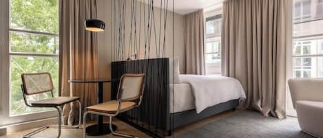 Une chambre d’hôtel moderne équipée d’un lit, de deux fauteuils, d’un bureau et de grandes fenêtres avec des rideaux.