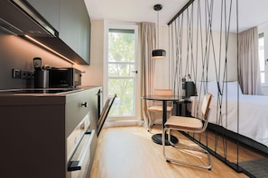 Ein modernes Hotelzimmer mit Küchenzeile, einer Essecke mit Tisch und Stühlen, einem großen Fenster mit Vorhängen und einem Bett mit schwarzem Kopfteil.