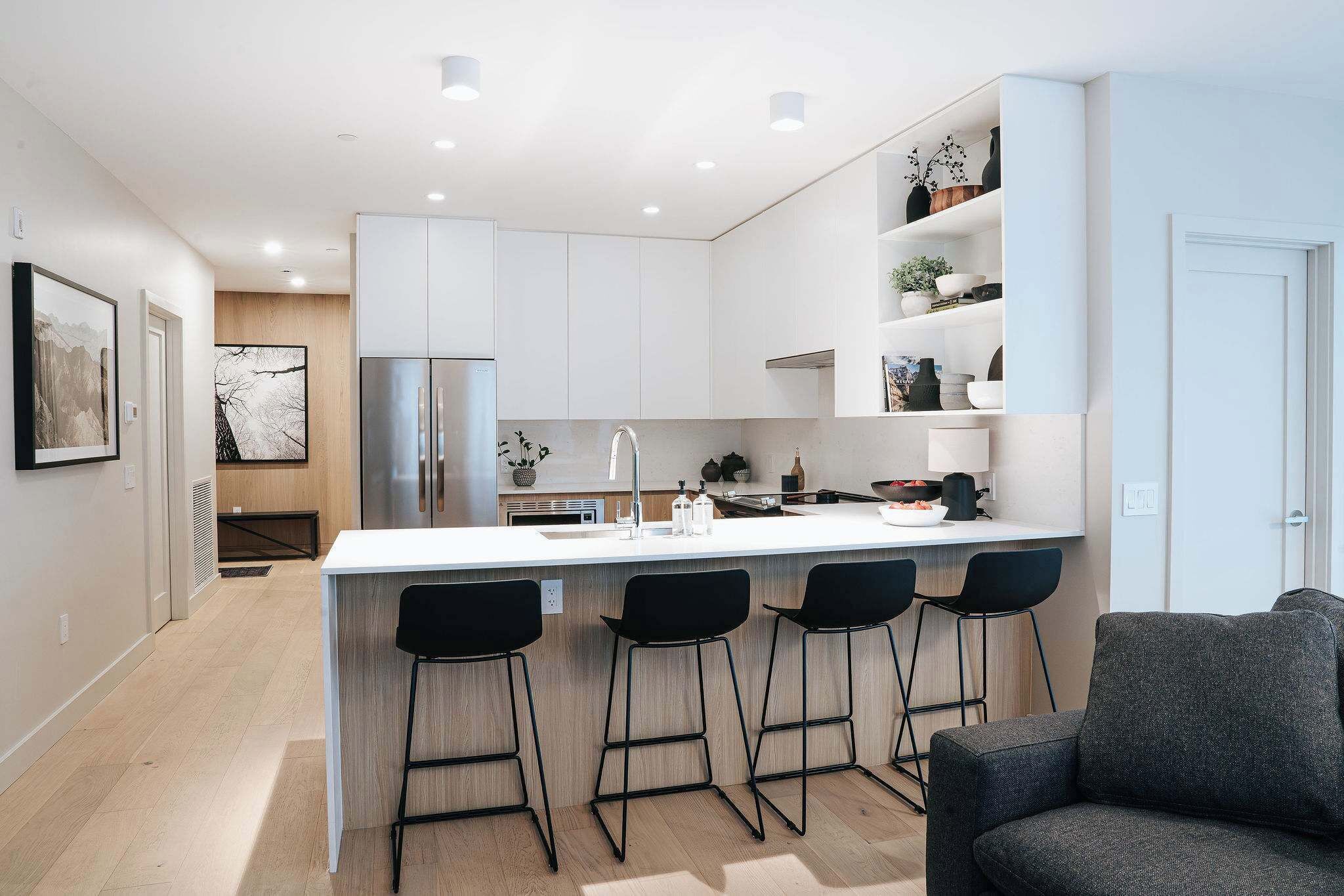 A modern kitchen with white cabinets, a stainless steel refrigerator, a central island with bar stools, and a grey sofa in the adjacent living area.