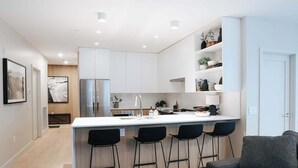 Een moderne keuken met witte kasten, een roestvrijstalen koelkast, een centraal keukenblok met barstoelen en een grijze bank in de aangrenzende woonkamer.