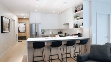A modern kitchen with white cabinets, a stainless steel refrigerator, a central island with bar stools, and a grey sofa in the adjacent living area.