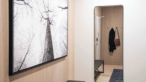 Een kamer met een groot schilderij van een boom, een houten garderobe, een bankje en een spiegel die een jas en tas reflecteert.