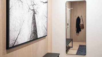 A room with a large framed picture of a tree, a wooden wardrobe, a bench, and a mirror reflecting a coat and a bag.