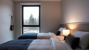 Een hotelkamer met twee bedden, een raam met uitzicht op bomen, een lampje op het nachtkastje en een kapstok met een tas.