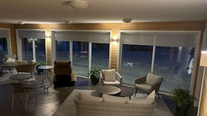Een moderne woonkamer met een bank, fauteuils, een salontafel en een eettafel. Er zijn grote ramen met zonwering, een plafondventilator en een kamerplant.